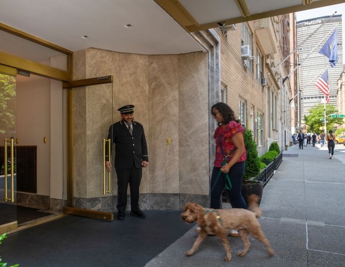 A doorman holds open a glass door for a woman and her small dog