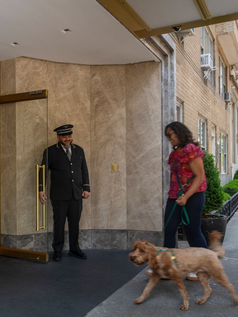 A doorman holds open a glass door for a woman and her small dog