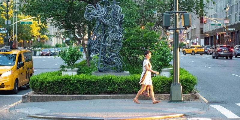 A woman crossing the street in front of a sculpture and trees in New York City