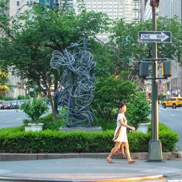 A woman crossing the street in front of a sculpture and trees in New York City