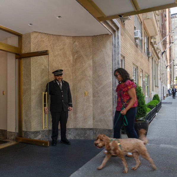 A doorman holds open a glass door for a woman and her small dog