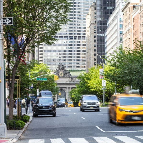 A busy New York City street showing a crosswalk with pedestrians, cars, and sidewalks lined with trees