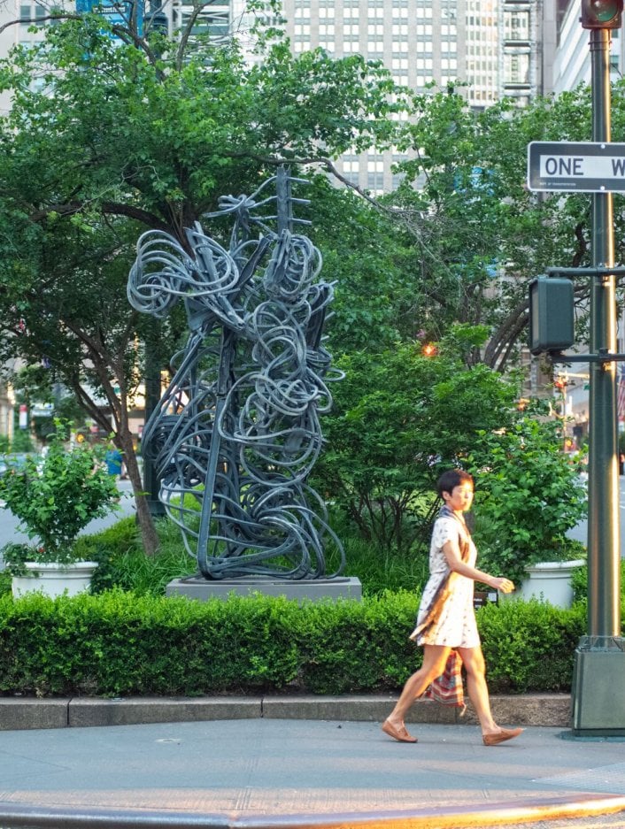 A woman crossing the street in front of a sculpture and trees in New York City