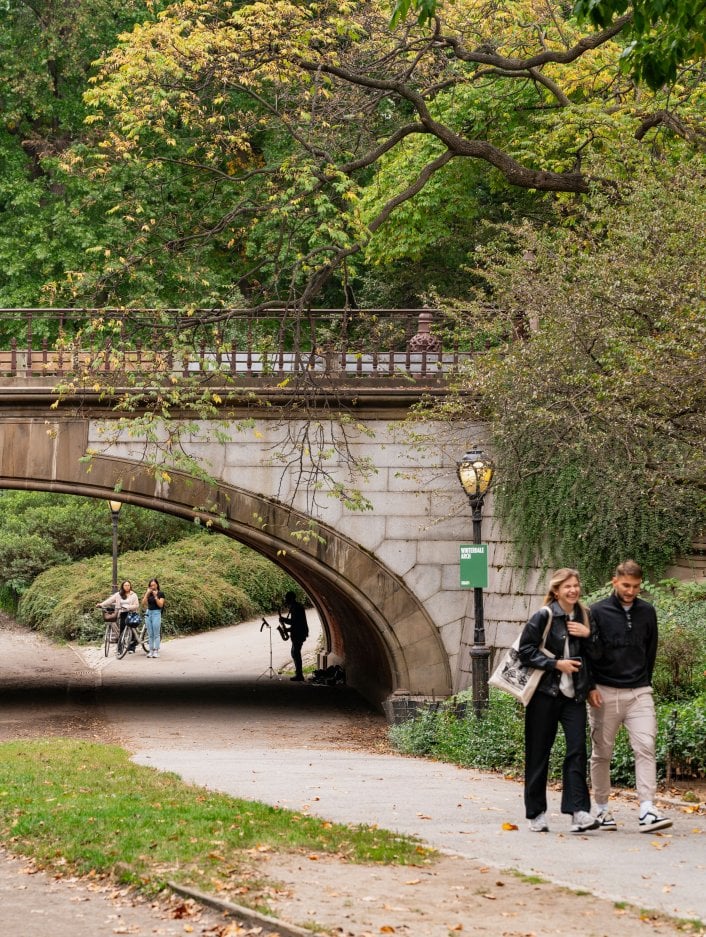 A couple walks along a path in Central Park with a bridge behind them